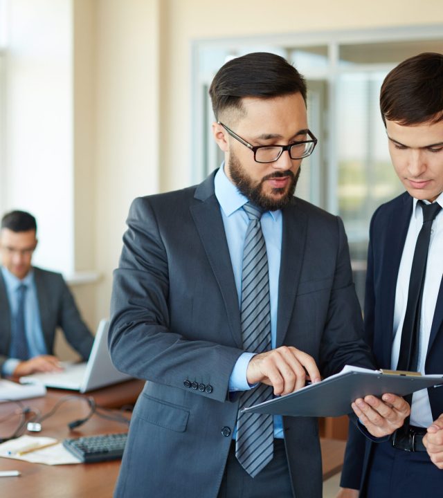 Confident businessman pointing at document while explaining his idea to his partner on background of their colleagues